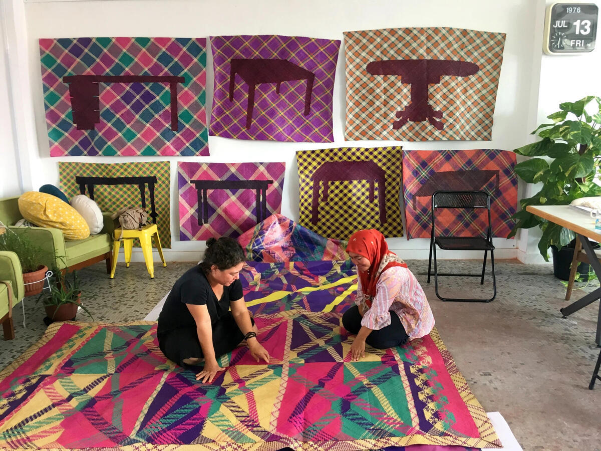 Two women sit on a colorful woven mat and converse. They are in a room with smaller mats that hang on the wall and contain motifs of tables.