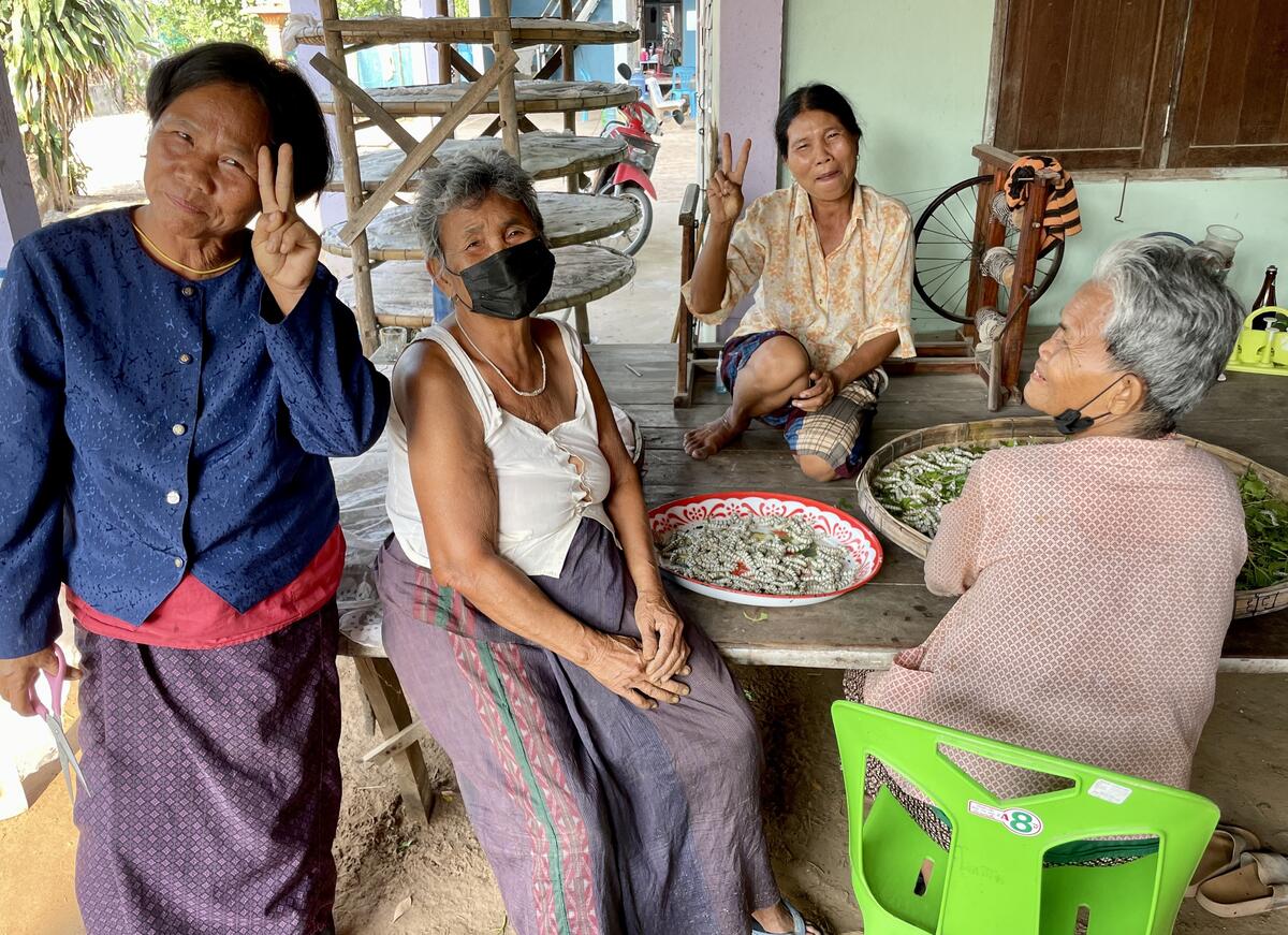 A group of four middle-aged and older women sit on and around a wooden platform and sort silkworms.